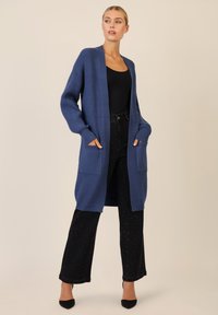 Pitkä sininen kardigan, joka on valmistettu ribbikuvioidusta neuleesta. Sen edessä on auki oleva malli, kaksi etutaskua ja rento hihansuun malli. Yhdistettynä mustiin housuihin.