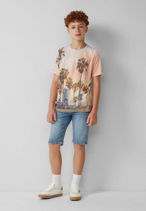 T-shirt grafica bianca con stampa di palme, pantaloni shorts in denim azzurro chiaro, e sneakers bianche con accenti color ocra, indossate con calze bianche.