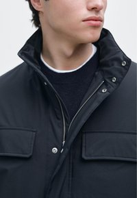 Zwarte jas met een hoge kraag, ritssluiting en twee zakken aan de voorkant. Heeft een gladde textuur en metalen drukknopen. Gedragen over een donkere trui.