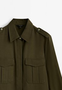 Olive green long-sleeve shirt with a collar, two front pockets, shoulder epaulets, and button accents on the shoulders. Smooth fabric texture.