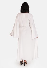 Robe maxi blanche avec de longues manches évasées. Tissu lisse et léger froncé à la taille. Portée avec des sandales à plateforme noires.
