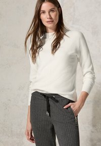 Pull en textile blanc avec col haut, associé à un pantalon gris foncé à rayures avec cordon de serrage et poches zippées.