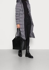 Abrigo acolchado gris, suéter de cuello alto negro, jeans oscuros, bolso tote de cuero negro y botas negras con cordones, superficie texturizada y suela gruesa.