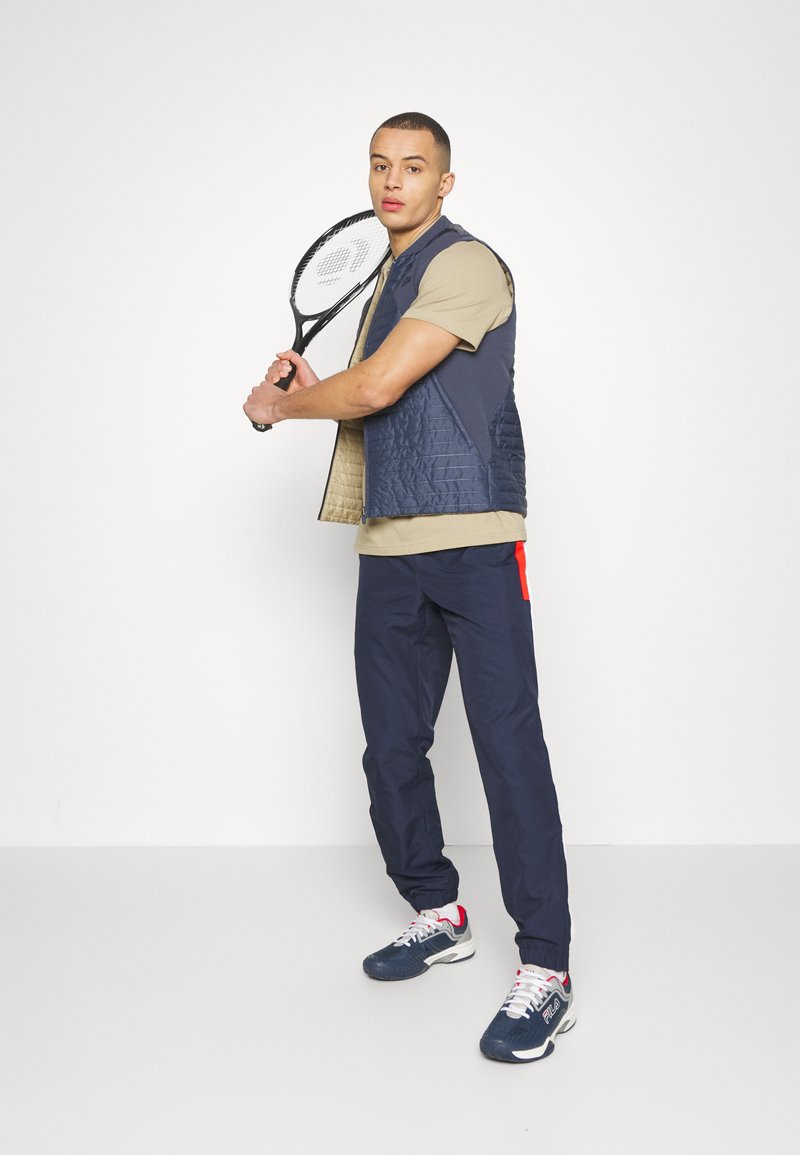 Gilet matelassé marine sur un t-shirt beige, associé à un pantalon marine et des chaussures marines avec des accents rouges, tenant une raquette de tennis.