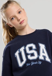 Navy blue sweatshirt made of cotton, featuring white "USA" text and "New York City" in light blue embroidery. Crew neckline and relaxed fit.