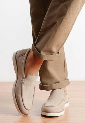 Chaussures beige en daim à enfiler portées avec un pantalon marron retroussé, posées sur un sol en bois clair.