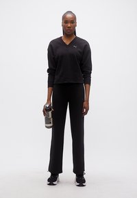 Γυναίκα που στέκεται και κρατά ένα διάφανο μπουκάλι νερού, ντυμένη με μαύρα αθλητικά ρούχα και αθλητικά παπούτσια, μπροστά σε απλό λευκό φόντο.