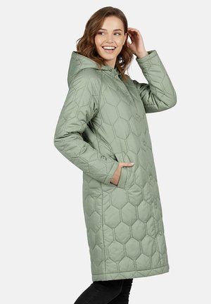 Vrouw die lacht terwijl ze een lange, lichtgroene jas met een zeshoekig ruitpatroon draagt, voorzien van een capuchon, verborgen knopen aan de voorkant en zijzakken.