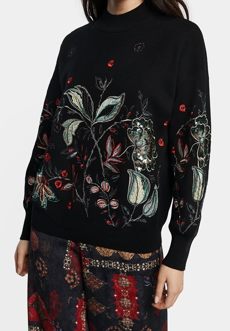Pull noir avec une broderie florale complexe en vert, rouge et beige, assorti à une jupe à motifs patchwork foncés.