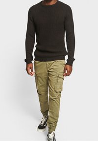 Pull noir en maille avec col rond, associé à un pantalon cargo vert olive avec poches latérales, et des baskets noires à lacets blancs.