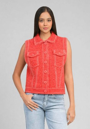 Femme portant un gilet sans manches corail à boutons avec poches frontales, associé à un jean bleu clair, debout devant un fond gris uni.