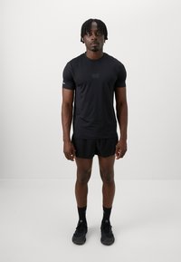 T-shirt de sport noir à manches courtes, avec un logo discret, associé à un short noir et des chaussures de sport noires. Fond uni.