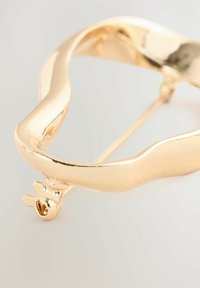 Guldtonet metalbroche med et bølget design. Har en nålelukning og en glat, skinnende overflade. Bemærkelsesværdig detalje inkluderer et unikt endestykke.