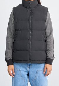 Schwarze Steppweste mit hohem Kragen, ausgestattet mit einem gesteppten Design, Reißverschluss vorne und seitlichen Taschen. Akzentuiert durch Ärmeln mit grauem Fischgrätenmuster.