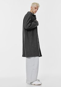 Szürke gyapjú keverék kabát laza szabással, elülső gombokkal és oldalsó zsebekkel, laza szürke sportnadrágok és fehér sportcipők felett.