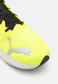 Zapatilla de running de malla amarilla brillante con cordones negros, que presenta un logotipo plateado texturizado y una suela blanca con un acento negro.
