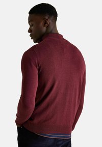 Vínovočervený pletený sveter s vysokým golierom, rebrovanými manžetami a lemom. Tkanina vyzerá mäkko s hladkým povrchom a bez viditeľných vzorov.