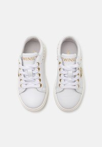 Sneakers in pelle bianca con occhielli e logo dorati. Punta arrotondata, suola piatta e borchie decorative sul tallone. Lacci in cotone, design a bassa altezza.