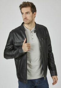 Zwarte leren jas met een staande kraag, ritszakken en geribde schouders. Draag het over een lichtgrijs shirt en blauwe jeans.