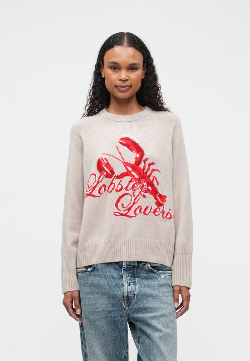 Mujer con pelo rizado y largo, vistiendo un suéter beige claro con un gran langosta roja y el texto "Amantes de los Langostas", combinado con unos jeans azules.