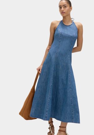 Kvinde iført blå denim halterkjole uden ærmer, der holder en brun taske, kombineret med brune snøresandaler med perledekorationer.