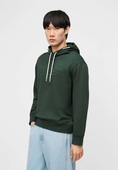 Νέος άνδρας ντυμένος με σκούρο πράσινο φούτερ με λευκά κορδόνια και ανοιχτό μπλε τζιν, στέκεται μπροστά σε ένα απλό ανοιχτό φόντο.