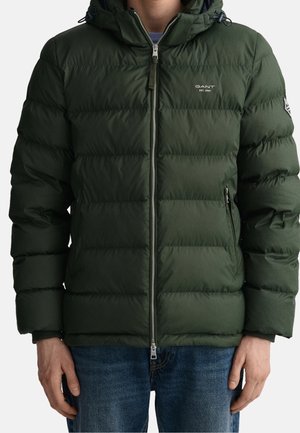 Man draagt een donkergroene doorgestikte pufferjas met een rits aan de voorkant, met rits afgesloten zakken en het GANT-logo op de borst en een patch op de mouw.