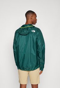Groene lichtgewicht jas met capuchon, voorzien van elastische manchetten en een trekkoord onderaan, versierd met een logo op de bovenrug, gladde textuur.