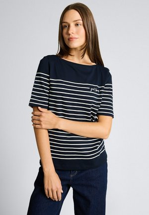 Kobieta o prostych brązowych włosach, ubrana w granatowo-biały paskowany krótki rękaw z napisem "good things", pozująca z założonymi ramionami.