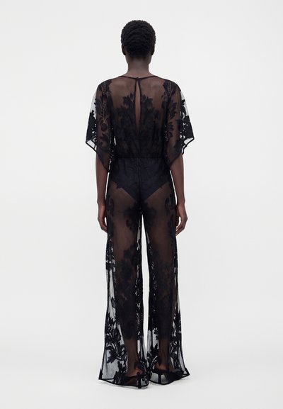 Femme vue de dos portant une combinaison noire en dentelle transparente avec des motifs floraux et des manches courtes sur un fond blanc uni.