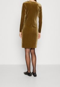 Robe en velours vert olive à manches longues, coupe droite, avec un détail de couture au dos. Portée avec des ballerines noires et des collants transparents.