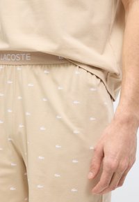 Μπεζ σορτς με μικρά λευκά λογότυπα κροκοδείλου και μπεζ ζώνη με λογότυπο, φορεμένα με αντίστοιχο μπεζ πουκάμισο, το χέρι ακουμπάει στο πλάι.