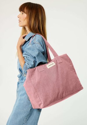 Rosa Tragetasche aus Stoff mit zwei kurzen Griffen. Auf einer Seite befindet sich ein Etikett. Über der Schulter zu einem Jeans-Outfit getragen.