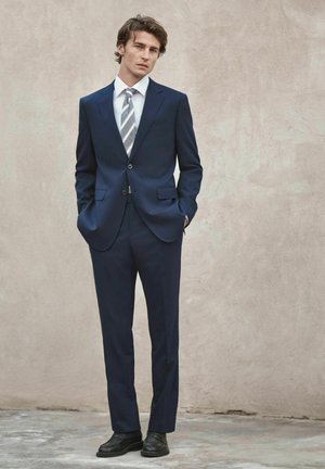 Jeune homme aux cheveux bouclés portant un costume bleu foncé, une chemise blanche, une cravate rayée, des chaussures noires, se tenant contre un mur texturé beige clair.