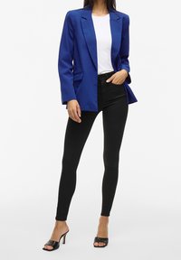 Femme portant un blazer bleu, un haut blanc, un pantalon skinny noir et des mules à talons noires ouvertes à l'avant, debout avec une main dans la poche.