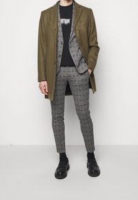 Manteau en laine vert olive porté sur un costume gris à carreaux avec des accents rouges. T-shirt noir et mocassins noirs complètent la tenue.