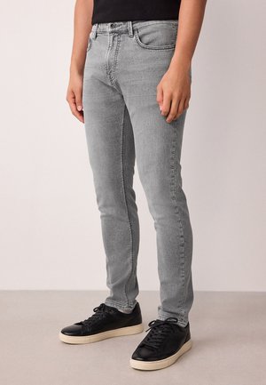 Persoon draagt lichtgrijze slim-fit jeans en zwarte sneakers met witte zolen, staande op een effen vloer.