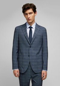 Gestructureerd blauw kostuum, enkel-breasted jas met notch revers, twee zakken, bijpassende broek, gecombineerd met een lichtblauw overhemd en een donker stropdas.