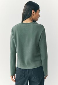 Maglione a coste verde oliva, con maniche lunghe, dotato di scollatura tonda e un design posteriore leggermente accorciato. Tessuto testurizzato con vestibilità aderente.