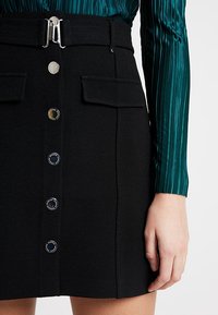 Mini-jupe noire avec poches avant, quincaillerie en argent, et une rangée de boutons circulaires noirs et argentés. Le tissu présente une finition texturée.