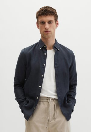 Giovane uomo con capelli castani corti che indossa una camicia blu navy abbottonata sopra una t-shirt bianca e pantaloni beige con coulisse, in piedi con le mani nelle tasche.