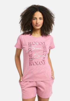 Vrouw met krullend haar die een roze T-shirt en bijpassende korte broek met tekstontwerp draagt, staand met handen in de zakken tegen een witte achtergrond.