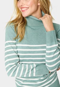 Gaiši turquoise džemperis ar ribotu augsto apkakli. Iezīmēts ar horizontālām baltām svītrām. Mīksta tekstūra un pieguļoša aproce, piemērots slāņošanai.