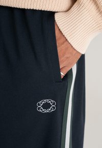Hand in de zak van een marineblauwe broek met een wit geborduurd logo en groen/witte zijstrepen, draagt een beige gebreide trui en een zilveren ring.