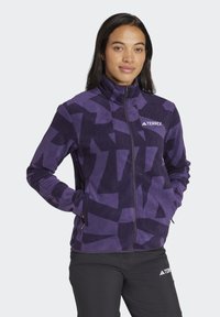 Chaqueta de forro polar púrpura con un patrón oscuro abstracto, cuello alto, cremallera frontal y bolsillos con cremallera, que presenta el logo de Terrex en blanco.