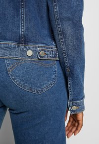 Jeansjacka i mörkblått med en texturerad yta; har silverknappar och en böjd fickdetalj på baksidan.