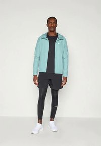 Light turquoise zip-up jacket with hood, worn over a black top and black shorts; black leggings and white sneakers complete the athletic look.