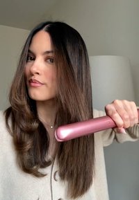 Prostownica do włosów o metalicznym różowym korpusie, trzymana w trakcie użycia, kontrastująca z ciemnymi, prostymi włosami, częściowo stylizowanymi z luźnymi lokami.