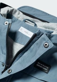 Vaaleansininen takki, jossa on vetoketjullinen etuosa, hopeiset yksityiskohdat ja harmaa sisävuori. Sisällä on etiketti "84 URHEILUTIETO".