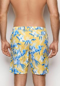 Costumi da bagno gialli con un motivo floreale e foglie blu. Presentano una vita elastica e due chiusure a bottone. Tessuto leggero.